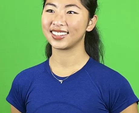 Photo of a young Asian woman smiling, with dark hair that is half up. She wears blue t-shirt and stands against a green background.