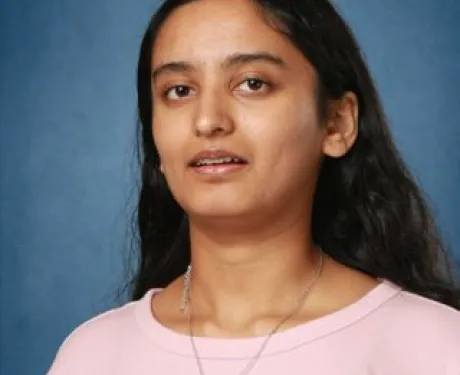 A young woman with brown skin and long dark brown hair looks at the camera. She is wearing a light pink top and a necklace with a red pendant..