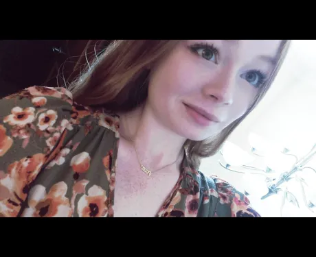 A young woman with brown hair and a flower shirt stands against a background that is brightly lit to the right and dark to the left.