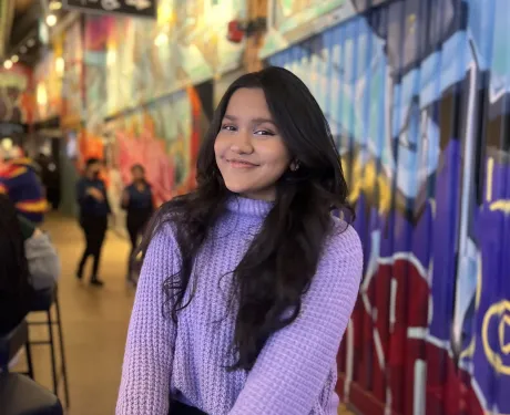 A young woman with brown skin and long dark hair wears a light purple sweater. She sits on a stool against a colourful graffiti background.