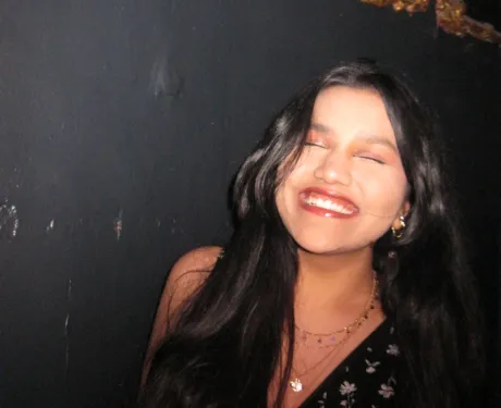 Photo of an Indian girl with long black hair smiling with her eyes closed against a dark background.