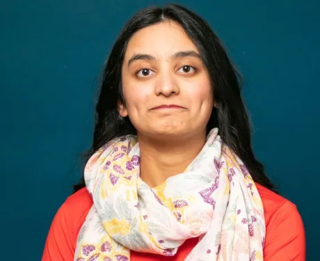 Photo of a young woman with light brown skin and long dark hair against a blue background. She wears a coral shirt and white scarf.
