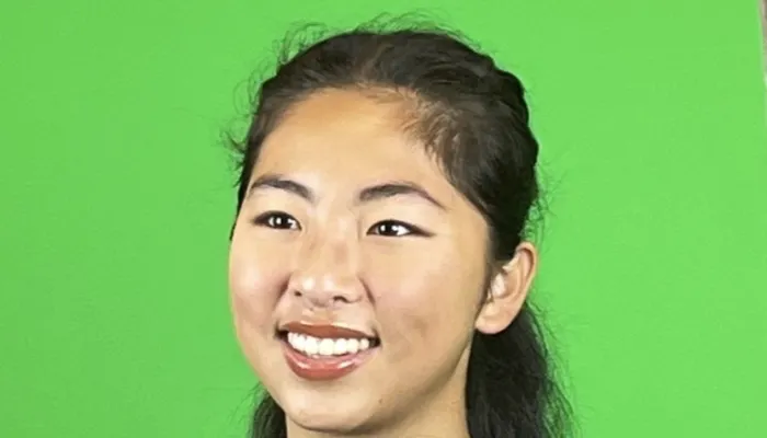 Photo of a young Asian woman smiling, with dark hair that is half up. She wears blue t-shirt and stands against a green background.