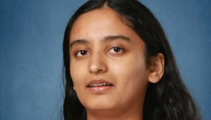 A young woman with brown skin and long dark brown hair looks at the camera. She is wearing a light pink top and a necklace with a red pendant..