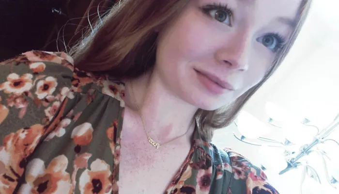 A young woman with brown hair and a flower shirt stands against a background that is brightly lit to the right and dark to the left.