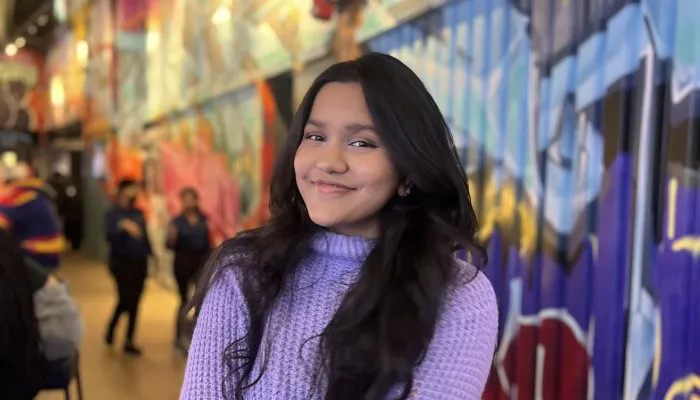A young woman with brown skin and long dark hair wears a light purple sweater. She sits on a stool against a colourful graffiti background.