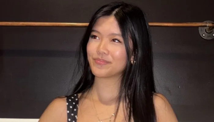 An asian girl with long black hair, wearing a polka dot tank top, stands with her head tilted to the left.