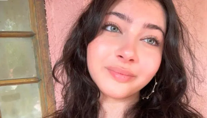 A Persian girl with blue eyes and long brown hair stands against a light pink wall and looks away from the camera. 
