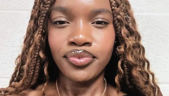 A young black girl stands against a white background. She is wearing a black one-shoulder top, a cross necklace, and has long hair with waves and braids in it.