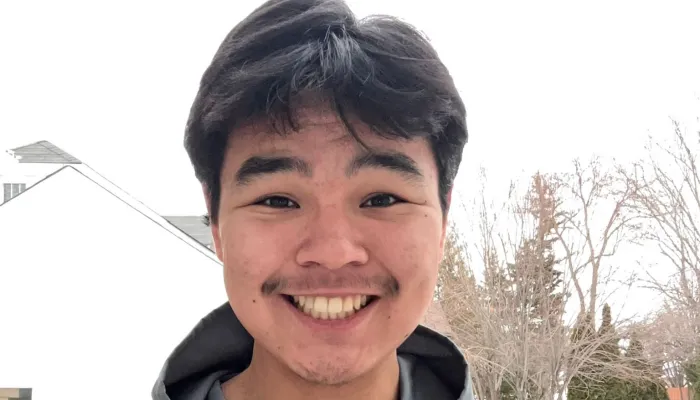 An Inuk teenage boy with short dark hair is smiling, standing outside against the snow and white sky.