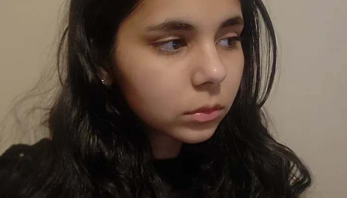 Shoulders-up photo of a girl with brown skin and long dark hair. She looks away from the camera and stands against a beige wall.