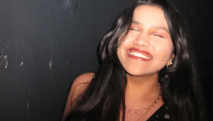 Photo of an Indian girl with long black hair smiling with her eyes closed against a dark background.