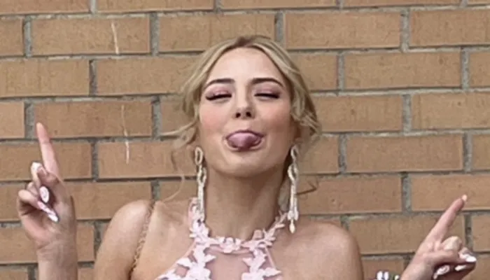 A girl in a formal pink dress sticks her tongue out at the camera and makes peace signs with her two hands. She stands against a brick wall.