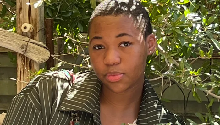 Photo of a black girl with a shaved head in an olive green jacket standing amongst tree branches and leaves. 