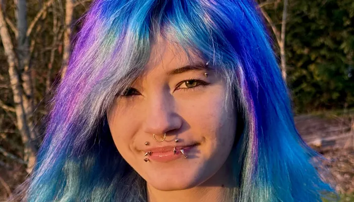 A girl stands outside against a backdrop of trees. She has many piercings and blue/purple hair.