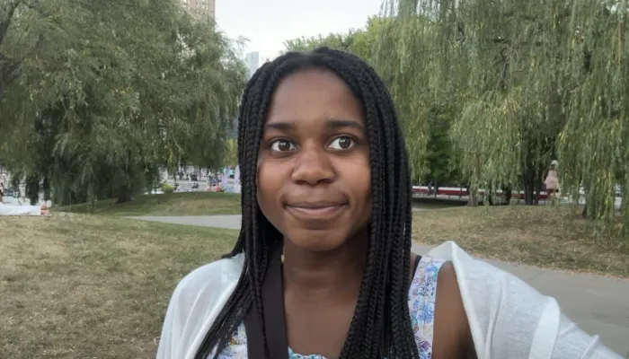 A black girl with long braids stands outside against a backdrop of trees.