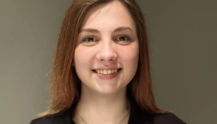 A teenage girl with light skin and brownish-reddish hair looks at the camera and smiles. She wears a short-sleeve black shirt with buttons and a collar.