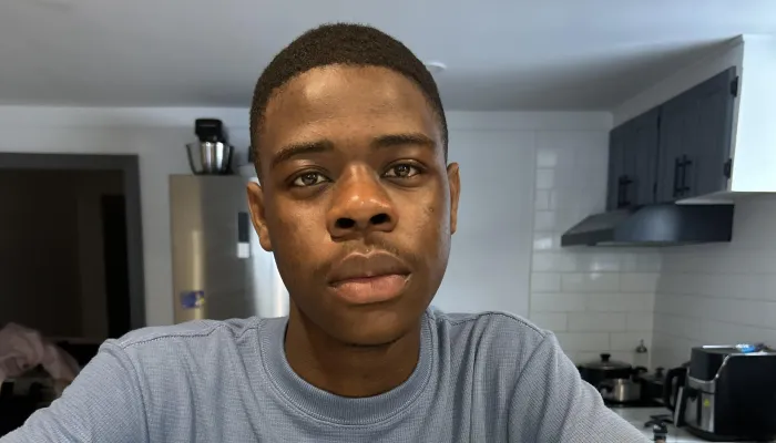 Photo of a young black boy in a light blue shirt staring directly at the camera. He is sitting at a counter with his arms resting on it. 