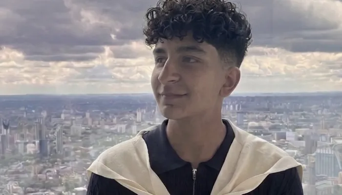 Photo of a young man with brown skin from the waist up in a dark tshirt with a white sweater wrapped around his shoulders. He stands against a city backdrop.