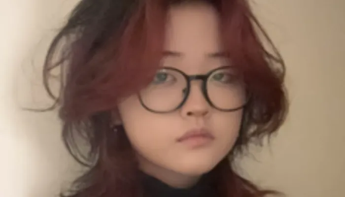 Photo of Chinese teenager with shoulder-length brown and reddish hair. They are wearing a dark shirt and large round glasses. 