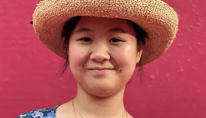 Photo of an Asian girl against a red wall. She wears a floral dress, two gold necklaces, and a straw hat over dark hair that is pulled back.
