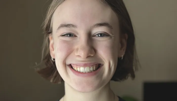 Headshot of a young white girl in an olive green shirt with a silver necklace. She has short brown hair and is smiling. 