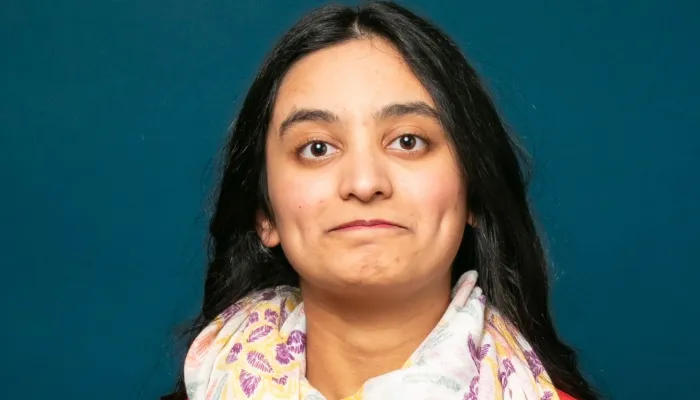 Photo of a young woman with light brown skin and long dark hair against a blue background. She wears a coral shirt and white scarf.